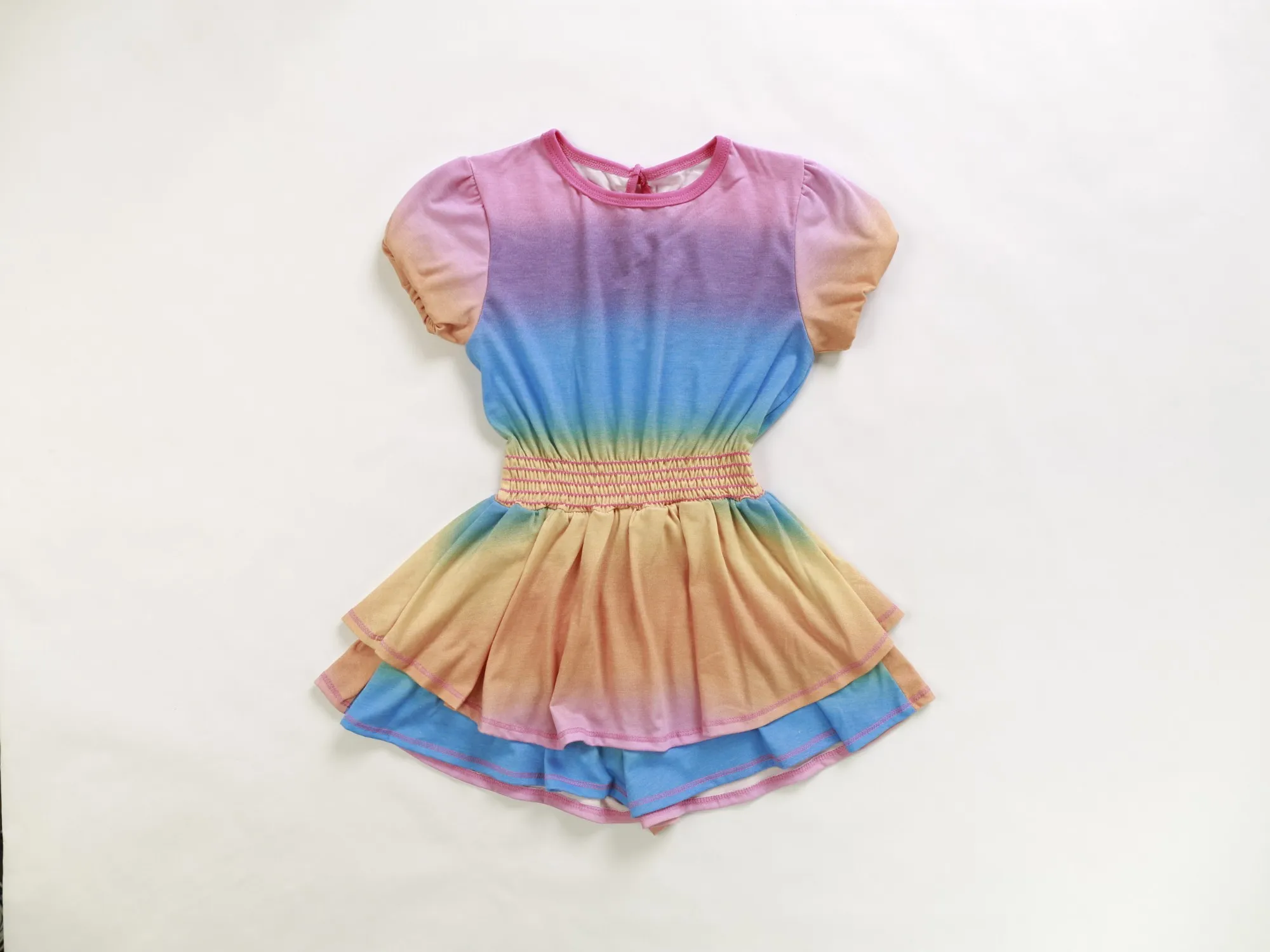 Rainbow ombré puff-sleeve dress laid on white paper, shot from overhead, with the paper's warm tone and a soft shadow around the garment
