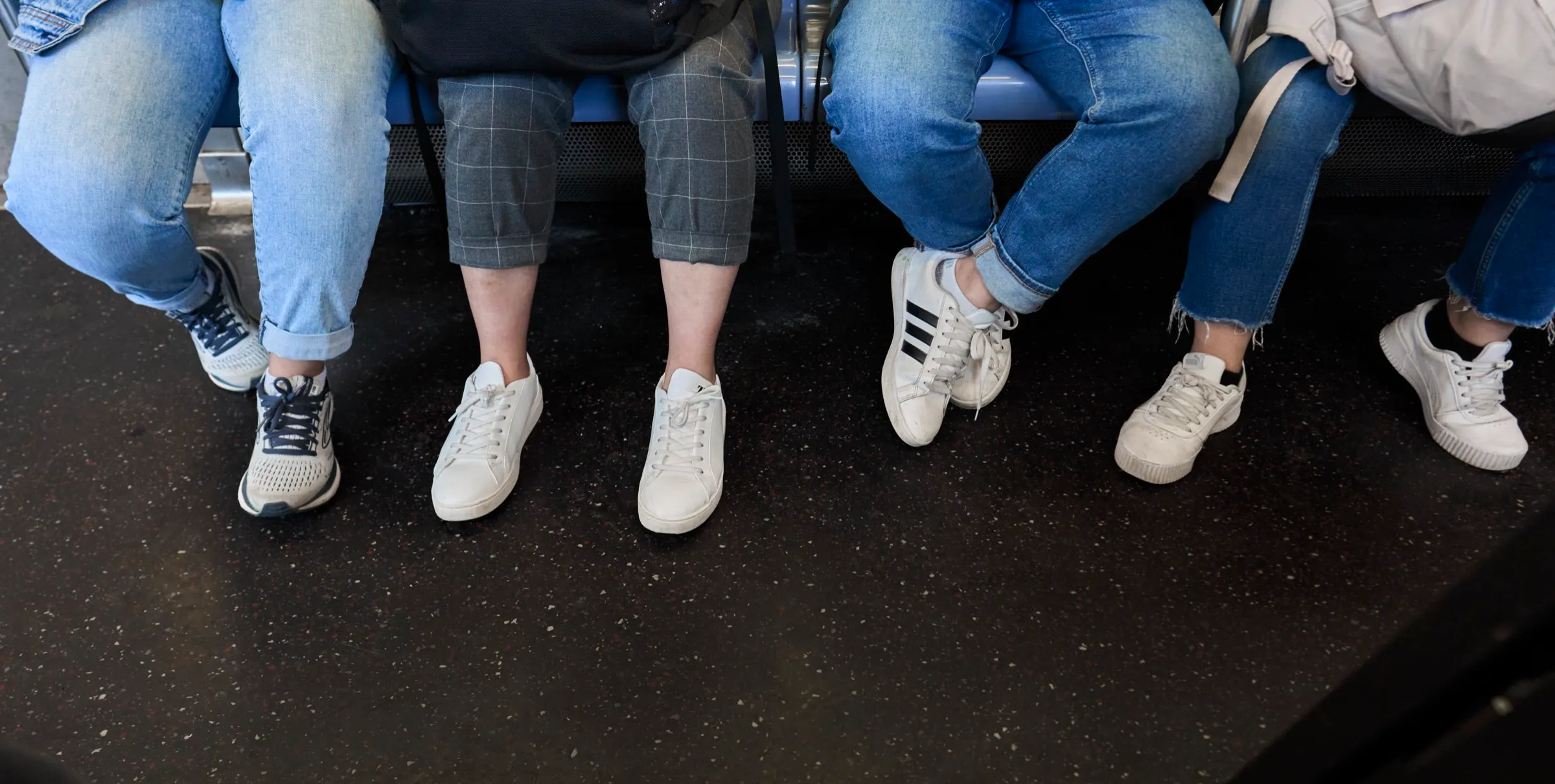 Four seated subway riders' legs in jeans and white sneakers, shot from across the aisle