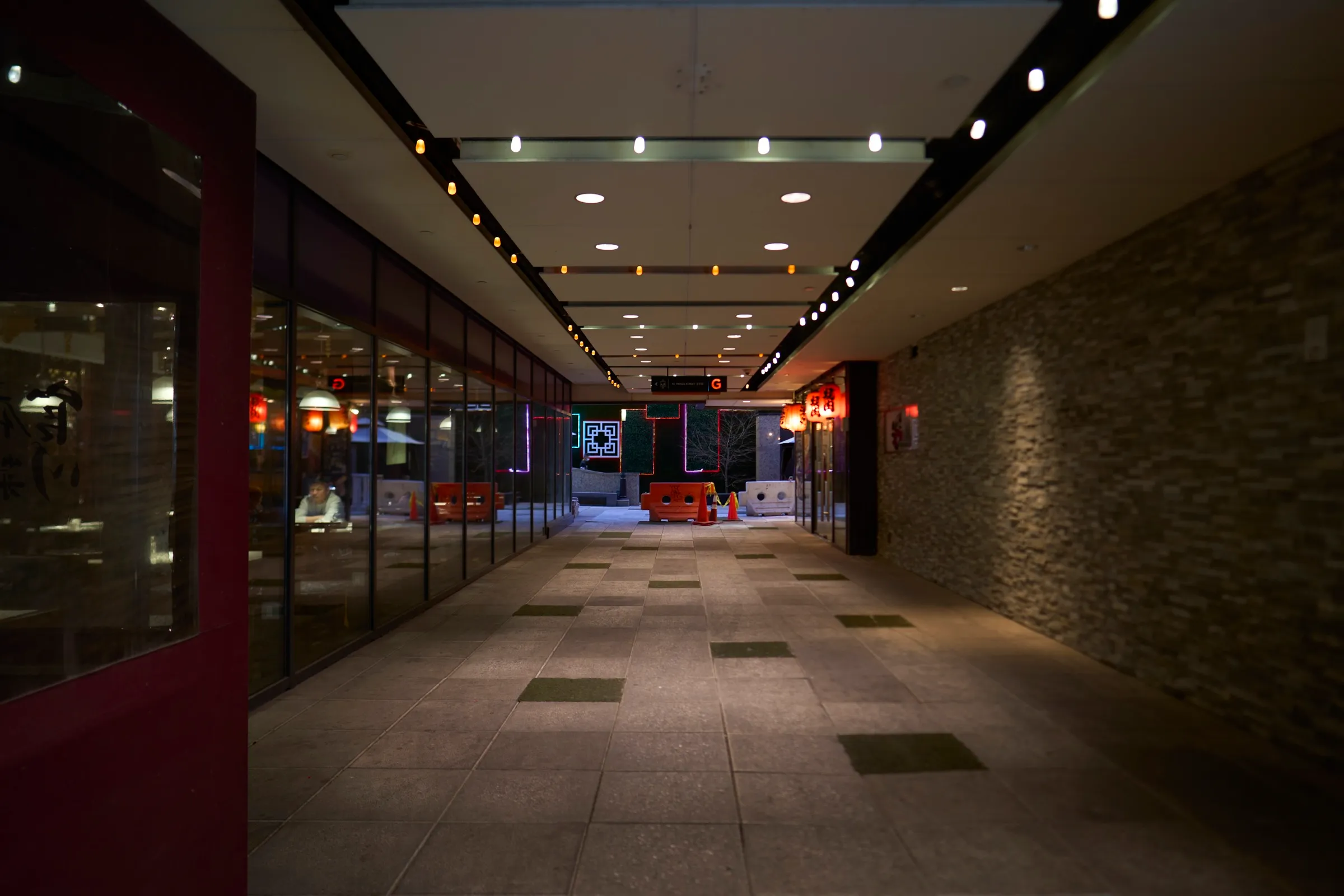 Evening walkway lit by rows of warm ceiling lights and neon signage