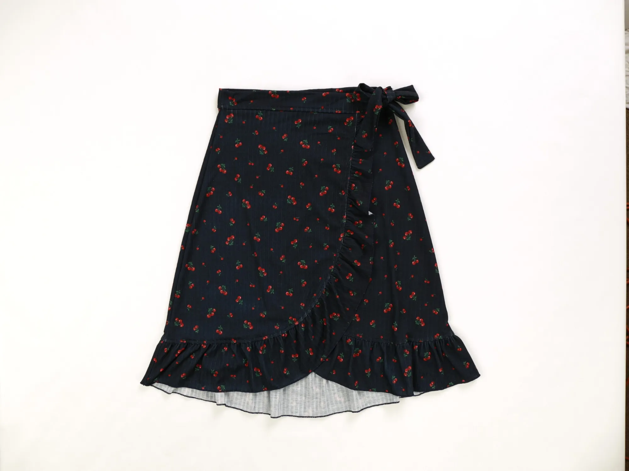 Navy cherry-print wrap skirt with ruffle hem and side tie, laid flat on white paper